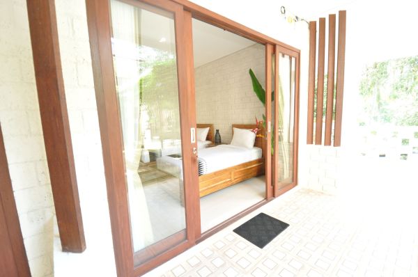 Bright tropical bedroom with open wooden sliding doors to a lush balcony, neatly made bed, and potted plants.